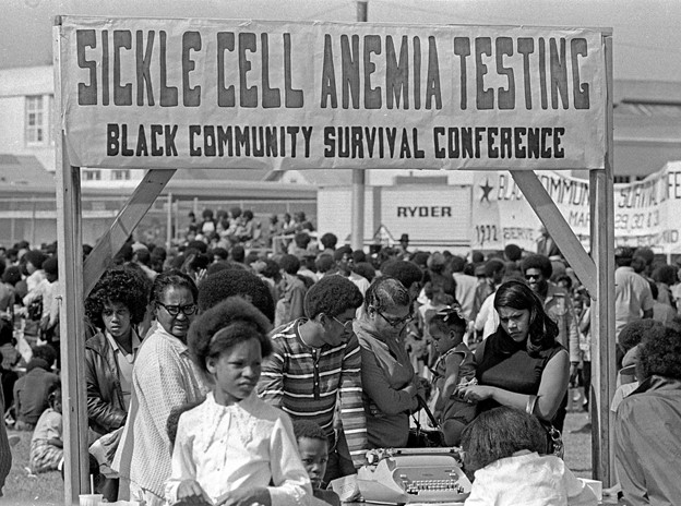 This black and white photograph depicts a crowd of people gathered outdoors. A large sign, held up by two wooden posts, is painted with the words “Sickle Cell Anemia Testing: Black Community Survival Conference” in the middle of the photograph. Beneath the sign is a large crowd of Black men, women, children, and elders sitting and standing outdoors.