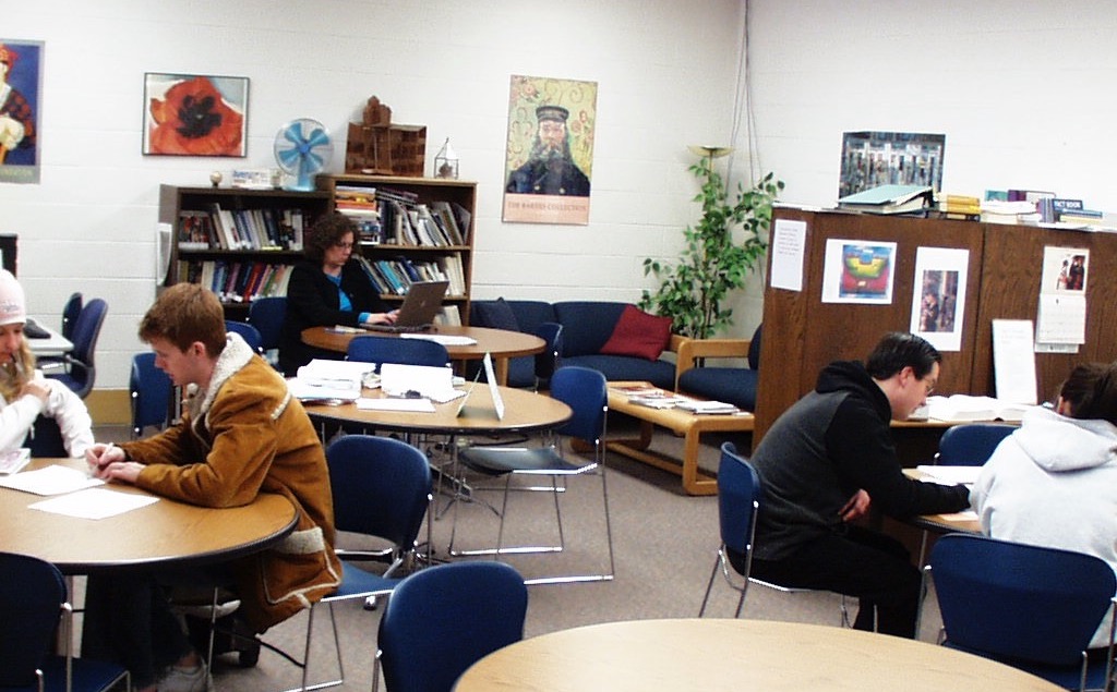 Photo of a the Salt Lake Community College Student Writing Center, which is a large room filled with wooden, circular tables. Each table has multiple chairs around them, and some of the tables are occupied by tutors and writers. There are two blue couches surrounded by bookshelves in the far back corner, and the walls are white with art hanging on them.